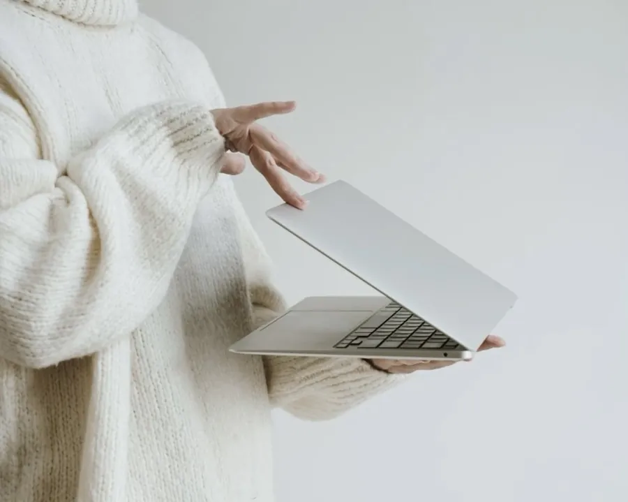 Person wearing a cozy oversized cream sweater holding a slim silver laptop in one hand while gently opening the screen with the other, against a minimal light background.