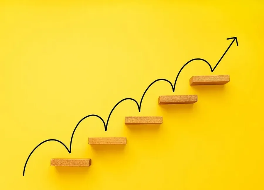 Stylized illustration of growth progression: wooden step blocks arranged upward on a bright yellow background with a curved line graph moving step-by-step upward and ending in an arrow pointing higher, symbolizing steady, structured growth.