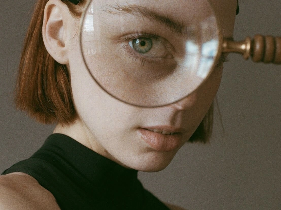 A close-up portrait of a person holding a magnifying glass over one eye, enlarging the green iris while the rest of the face remains softly in focus against a neutral background.