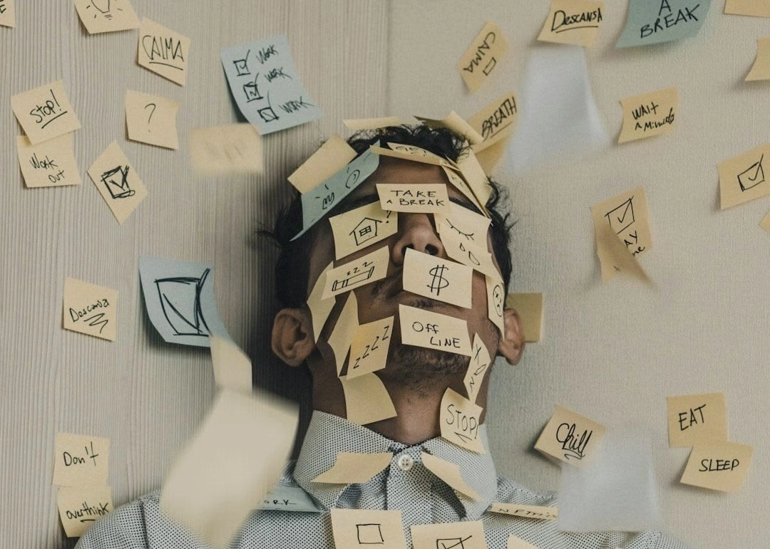 A person sits against a wall with their face covered in sticky notes while more notes float around them. The notes contain reminders like “stop,” “take a break,” “eat,” and “sleep,” creating a visual sense of stress, mental overload, and too many tasks at once.