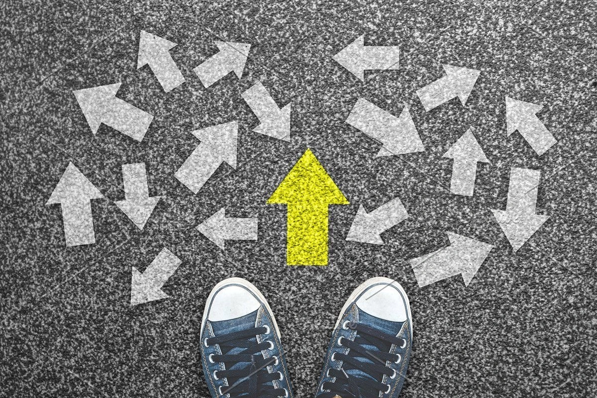 A person wearing sneakers stands on a gray surface covered with many white arrows pointing in different directions, while one bright yellow arrow points straight ahead, symbolizing choosing a clear direction among many options.