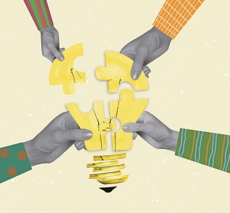 Four hands holding separate yellow puzzle pieces that together form a light bulb, symbolizing collaboration, shared ideas, and problem-solving.