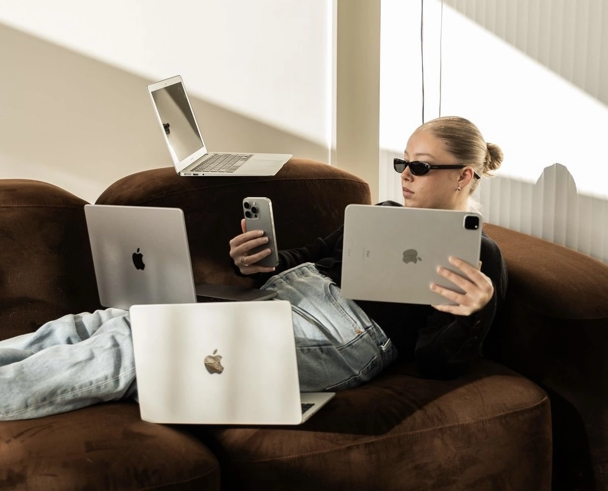 A person relaxing on a couch while multitasking with several Apple devices, holding a tablet and a smartphone while multiple laptops rest around them, suggesting digital work, productivity, or managing multiple online tasks at once.