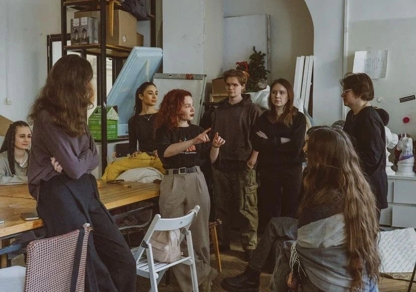 A group of people gathered in a creative workspace, standing and sitting around a table while one person speaks and gestures, suggesting a team discussion or collaborative brainstorming session.
