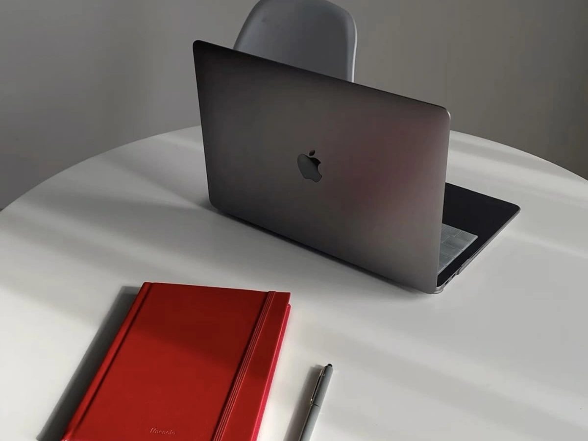 A minimalist workspace with a silver laptop on a white table, alongside a bright red notebook and a pen, set under soft natural lighting.