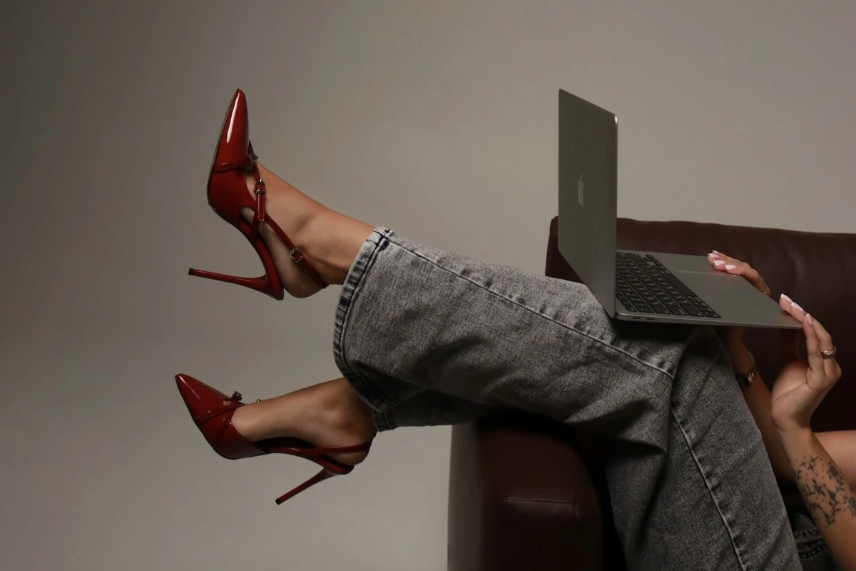 A person sitting on a chair with their legs crossed and resting on an armrest, wearing red high heels and jeans while using a laptop on their lap.