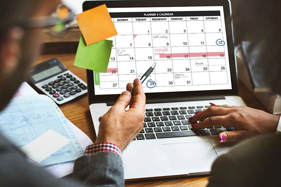 A person using a laptop to view a monthly calendar planner, pointing at specific dates with a pen, with sticky notes attached to the screen and a calculator and papers on the desk nearby.