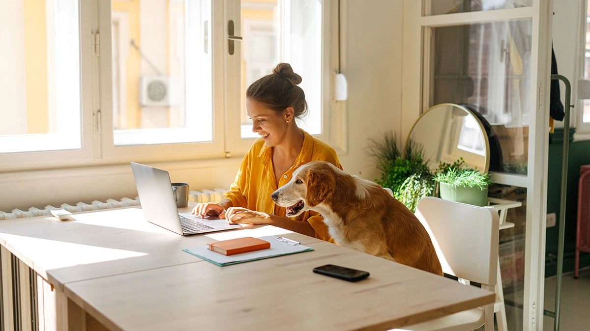 A woman working on a laptop at a bright home desk while a dog sits beside her, creating a warm and balanced work-from-home atmosphere.