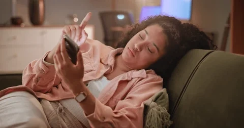 A woman lying on a couch, looking at her phone with a relaxed but slightly tired expression, illuminated by soft indoor lighting, suggesting late-night scrolling or unwinding after a long day.