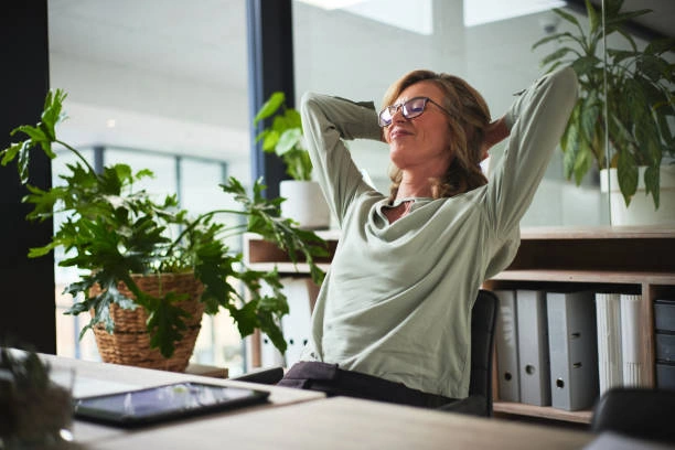 A relaxed woman leaning back in her office chair with hands behind her head, surrounded by plants and folders, enjoying a moment of calm and satisfaction at work.