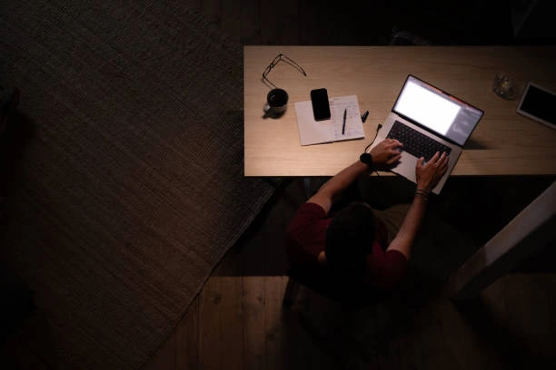 A man working late at night in a dark room, illuminated by the glow of a laptop screen, focused on his work while surrounded by a quiet, low-light environment that suggests concentration, fatigue, and long hours.