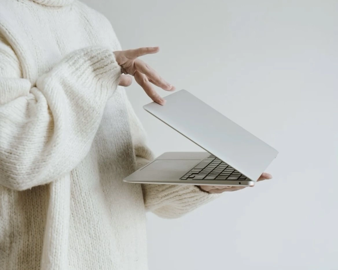 A person wearing a cozy oversized sweater holding a partially open laptop with one hand, while the other hand gestures above the keyboard, set against a clean, minimal background.