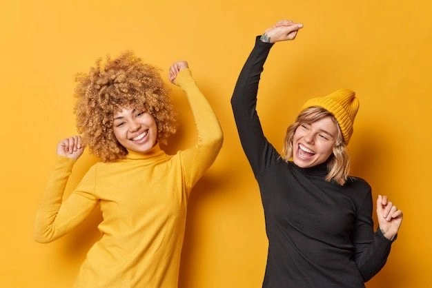 Two women smiling and dancing against a bright yellow background, expressing joy and energy, one wearing a yellow top and the other in black with a matching yellow beanie.