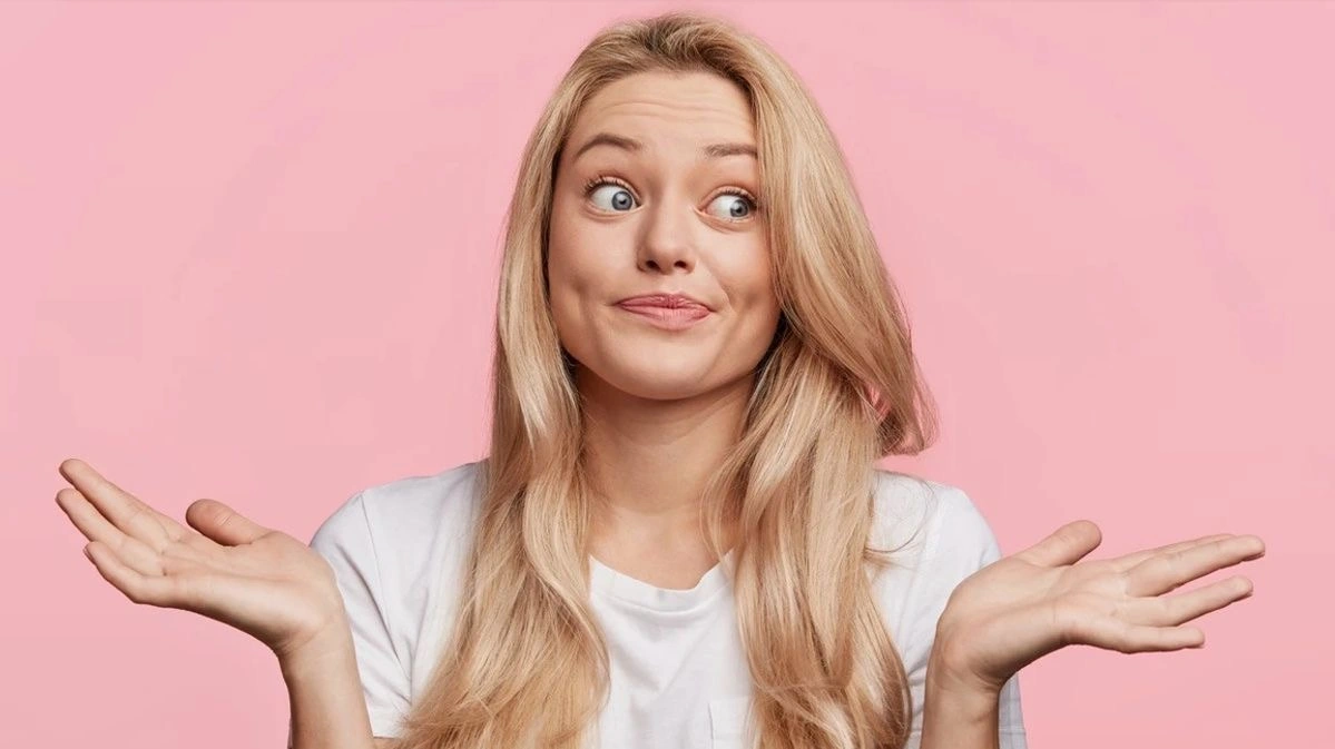 A young woman against a pink background shrugging with both hands raised, making a playful, uncertain facial expression as if saying “I don’t know.”