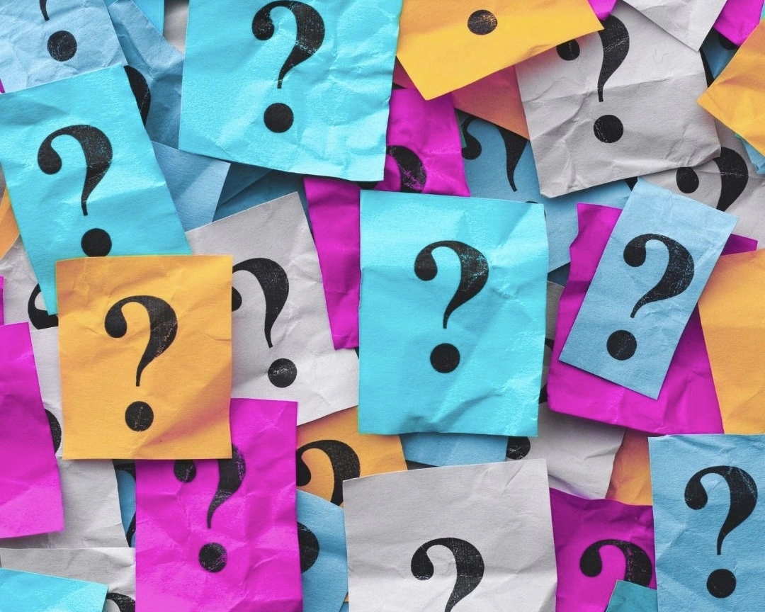 A colorful arrangement of overlapping sticky notes in bright blue, pink, yellow, and white, each marked with a black question mark, symbolizing curiosity, confusion, or brainstorming ideas.