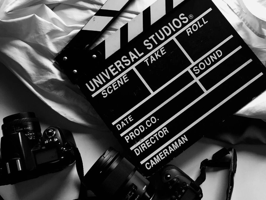 A black-and-white composition featuring a film clapperboard labeled “Universal Studios” placed on fabric, surrounded by professional camera equipment, symbolizing filmmaking, production, and behind-the-scenes creativity.