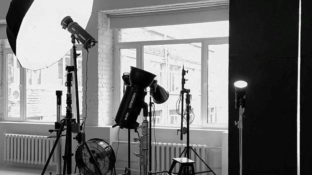 A black-and-white photo studio setup with professional lighting equipment, including softboxes, studio lights, and stands arranged near large windows, creating a minimal and industrial creative workspace.