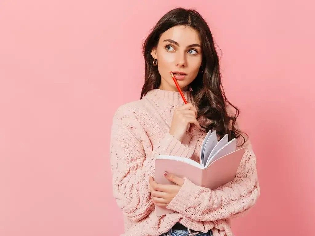 A young woman holding a notebook and pencil, looking to the side with a thoughtful expression against a soft pink background, as if brainstorming ideas or planning something.