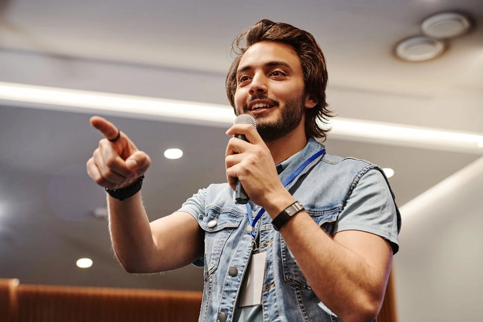 A charismatic young man with facial hair speaking into a microphone indoors, wearing a denim vest.