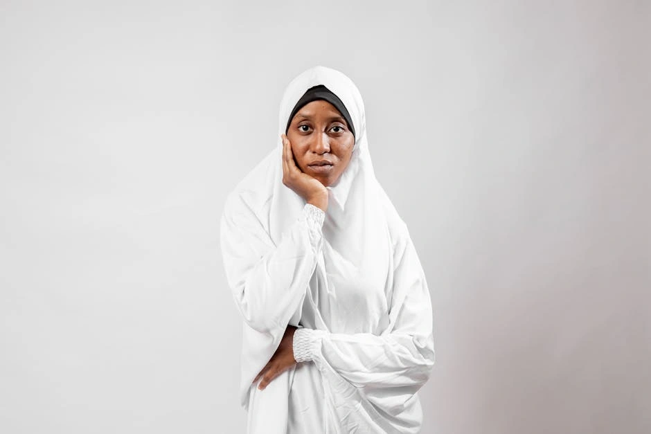 A portrait of a thoughtful woman wearing a traditional white hijab against a plain background.
