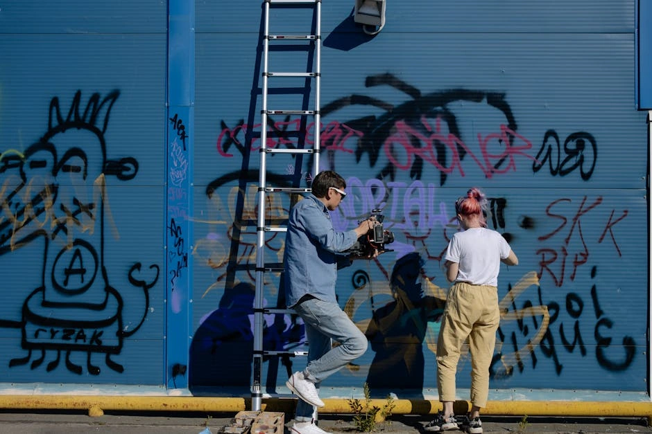 A man films a woman spray painting graffiti on a blue urban wall, showcasing street art.