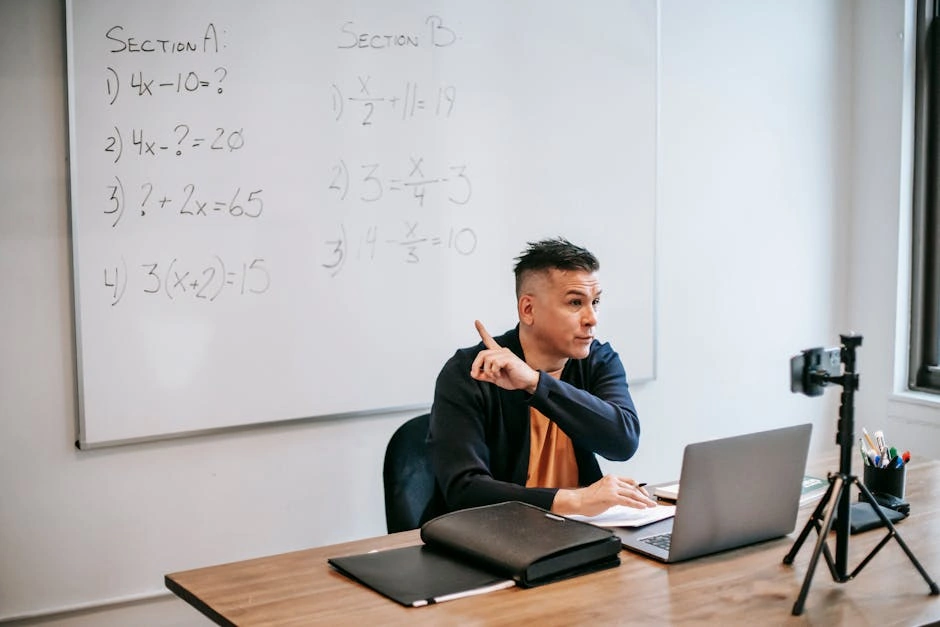 A male teacher explains math equations during an online class, using a laptop and whiteboard.